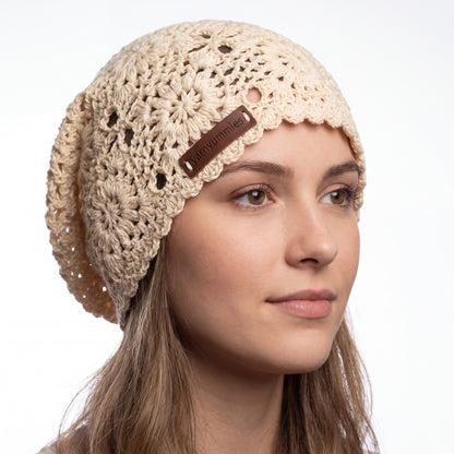 A woman wearing a beige crocheted slouchy beanie with a brown leather brand patch that reads 'yarnyummies'. The photo is taken against a clean white background.