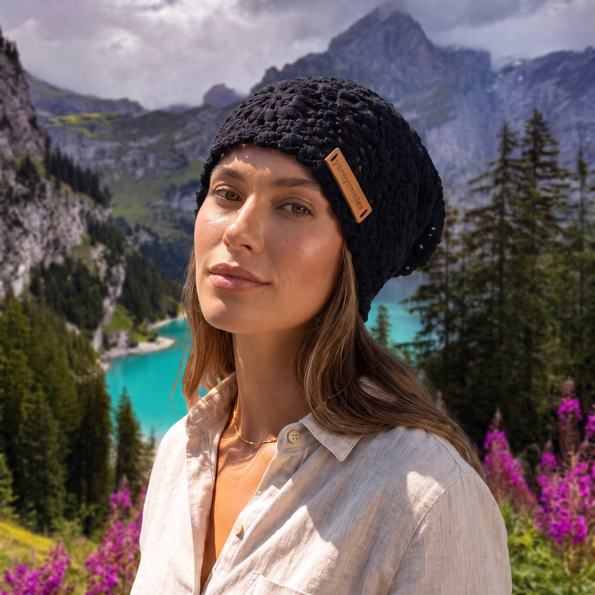 A woman wearing a black handmade crochet slouchy beanie by YarnYummies, posed outdoors in front of a scenic mountain and lake landscape.