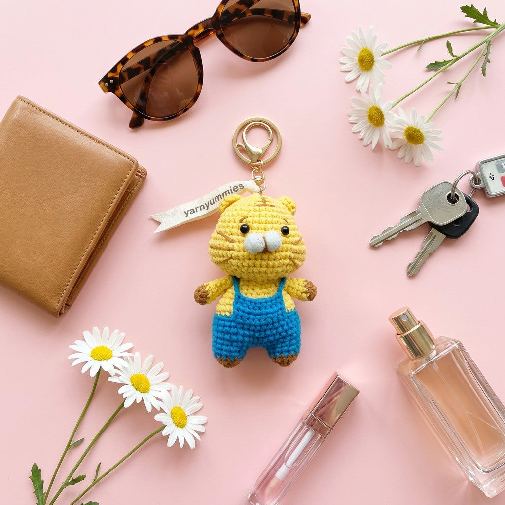 Crocheted keychain of a bear in overalls on a pink background with wallet, sunglasses, keys, and flowers.