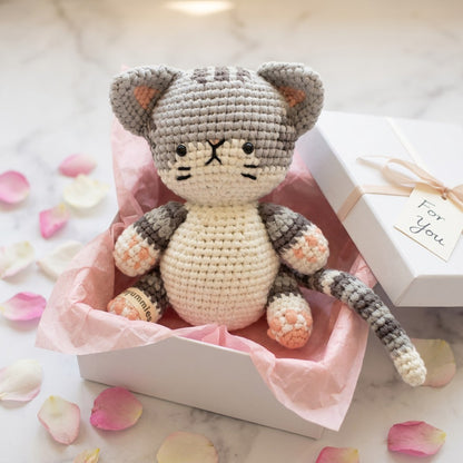 Crocheted cat toy in a white box with pink tissue paper, surrounded by rose petals.
