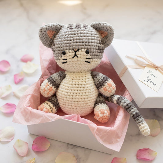 Crocheted cat toy in a white box with pink tissue paper, surrounded by rose petals.