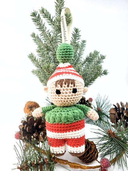 Handmade amigurumi elf with stitched eyes, wearing a red and green outfit, displayed among pine cones and branches.