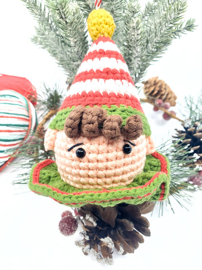 Handmade cotton yarn elf head ornament with a colorful striped hat, green and red features, and a rattle inside, displayed against a Christmas tree backdrop.