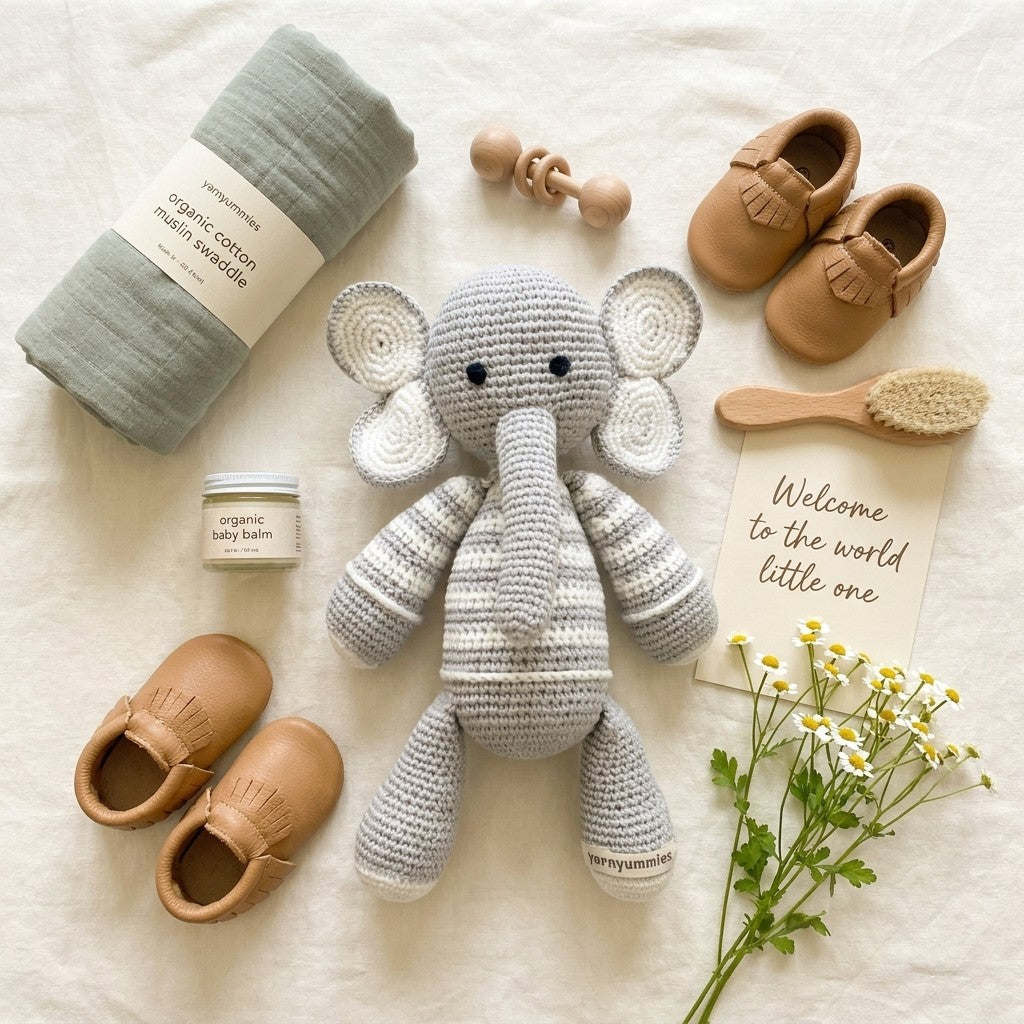Baby items including a crochet elephant toy, shoes, a blanket, and a card on a soft surface.
