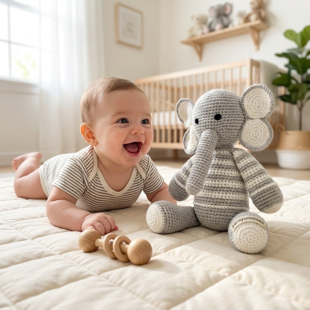 Baby lying on a bed with a crocheted elephant toy and wooden rattle in a nursery setting.