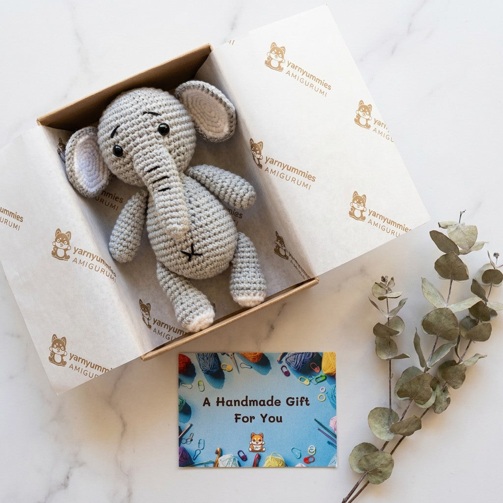 Crocheted elephant toy in a branded box with a card and eucalyptus leaves on a white background