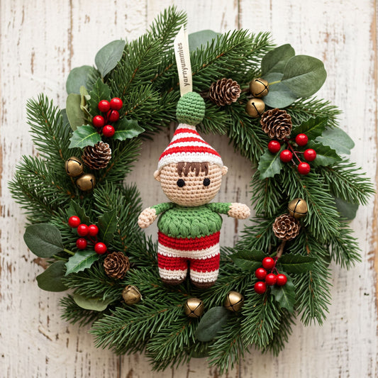 Handmade crochet baby safe Christmas elf ornament by YarnYummies hanging from a green pine wreath decorated with red berries, pinecones, and small gold bells, set against a whitewashed wooden background.