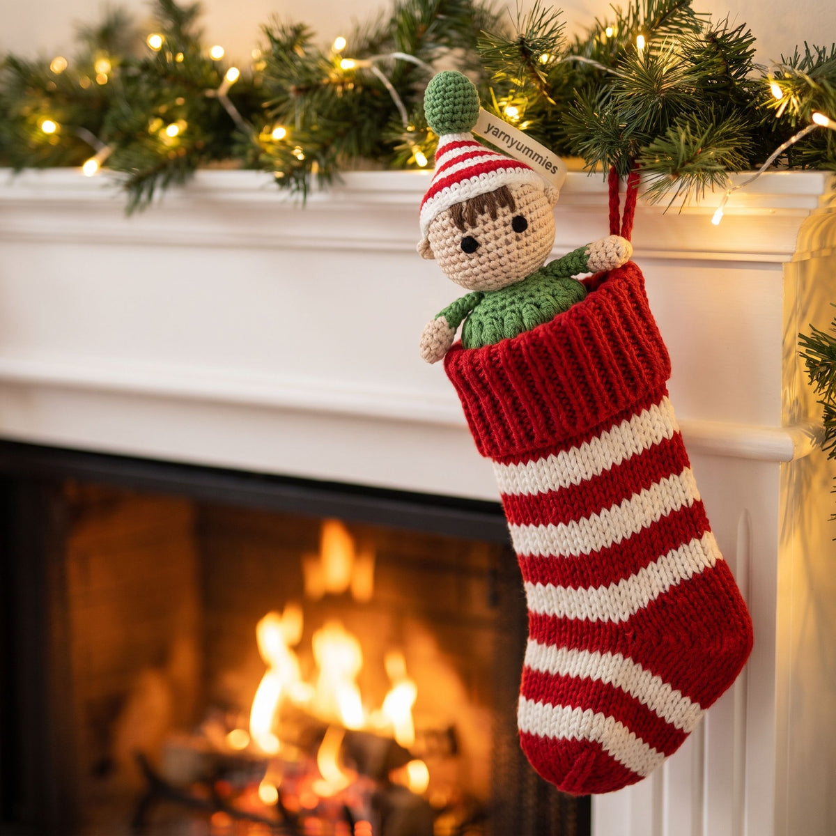 A red and white striped knitted Christmas stocking with a handmade crochet baby safe Christmas elf ornament by YarnYummies showing embroidered eyes and red and green outfit hanging from the opening, set against a cozy fireplace with a green garland and warm lights in the background.