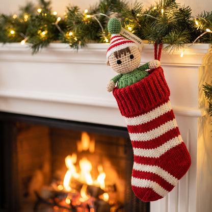 A red and white striped knitted Christmas stocking with a handmade crochet baby safe Christmas elf ornament by YarnYummies showing embroidered eyes and red and green outfit hanging from the opening, set against a cozy fireplace with a green garland and warm lights in the background.