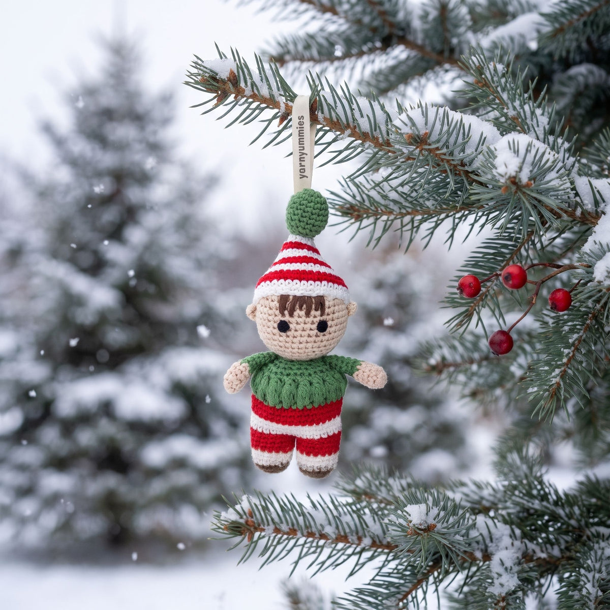 A handmade crochet baby safe Christmas elf ornament by YarnYummies showing embroidered eyes and red and green outfit hanging from a snowy evergreen tree branch. The ornament features a beige face and hands, a red and white striped hat with a green pom-pom, a green sweater, and red and white striped pants.
