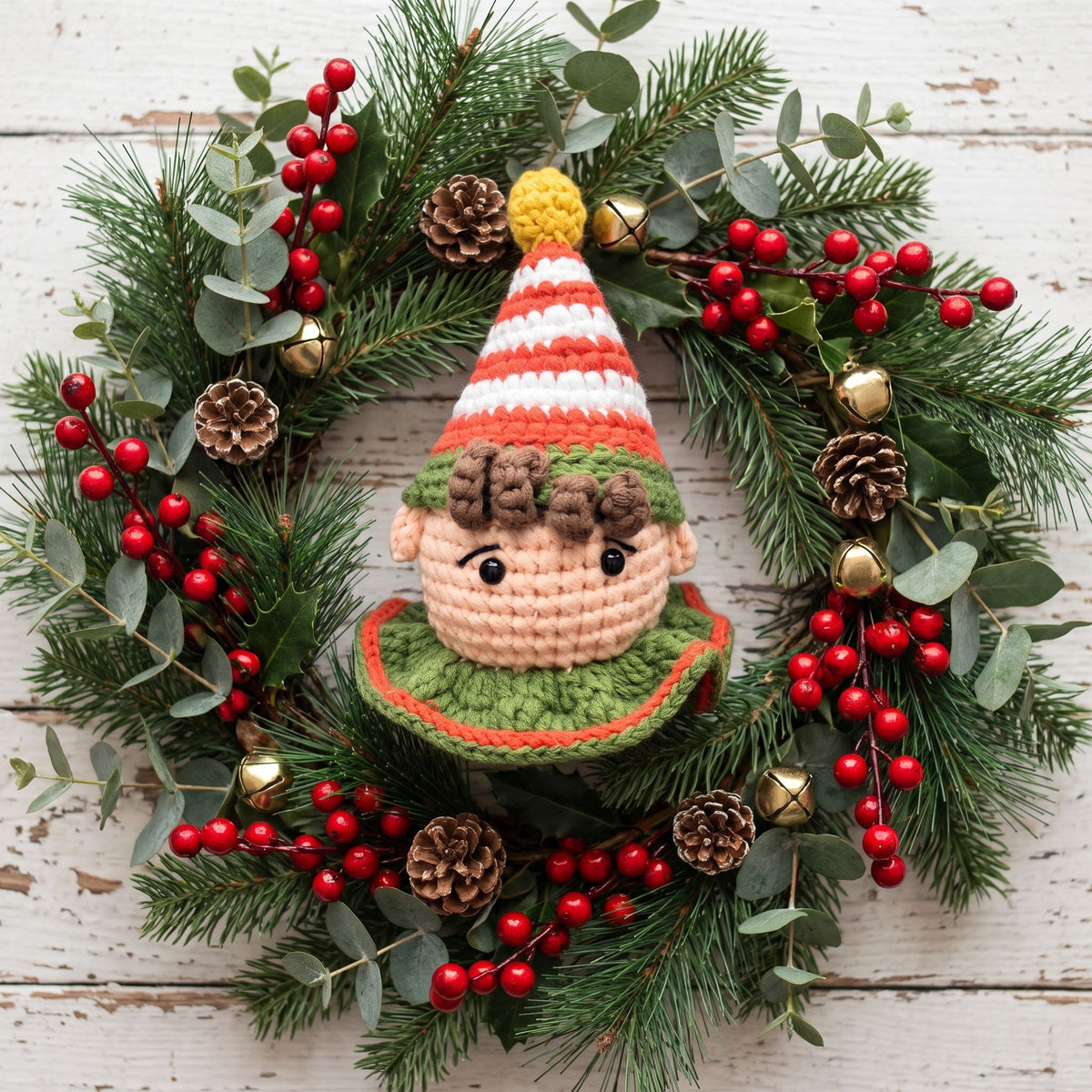 A festive Christmas wreath featuring a central Handmade crochet elf head ornament with rattle by YarnYummies front view showing rosy cheeks and wearing a striped red and white hat with a yellow pom-pom, surrounded by green pine branches, eucalyptus leaves, red berry garlands, pinecones, and small gold bells on a whitewashed wooden background.