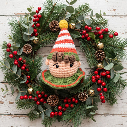 A festive Christmas wreath featuring a central Handmade crochet elf head ornament with rattle by YarnYummies front view showing rosy cheeks and wearing a striped red and white hat with a yellow pom-pom, surrounded by green pine branches, eucalyptus leaves, red berry garlands, pinecones, and small gold bells on a whitewashed wooden background.