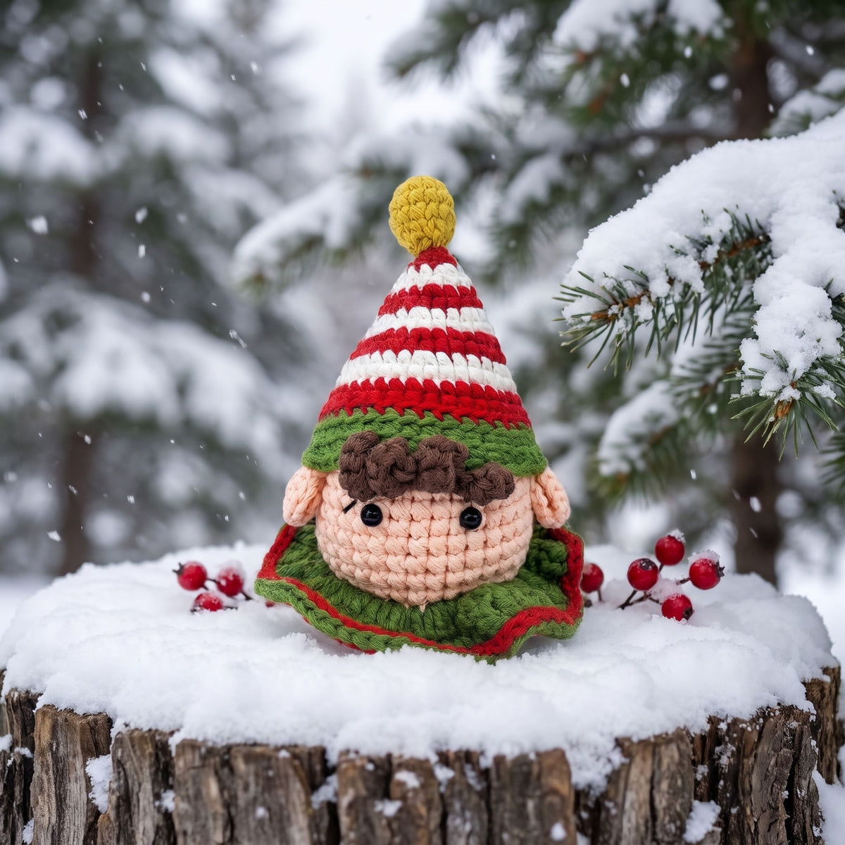 A Handmade crochet elf head ornament with rattle by YarnYummies front view showing rosy cheeks with a red and white striped hat, brown hair, and a green collar sits on a snowy log in a winter forest setting with snow-covered pine branches and falling snow.