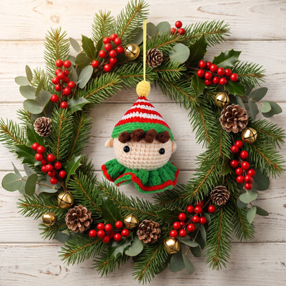 A festive Christmas wreath made of green pine branches and eucalyptus leaves, decorated with red berry clusters, pinecones, small gold bells, and a Handmade crochet elf head ornament by YarnYummies front view showing vibrant red and green striped hat hanging in the center against a whitewashed wood background.