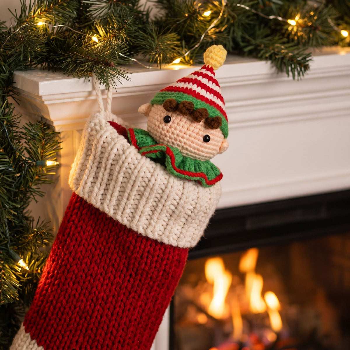 A knitted Christmas stocking with a Handmade crochet elf head ornament by YarnYummies front view showing vibrant red and green striped hat out, hanging from a white fireplace mantel decorated with green pine garlands and warm string lights, with a fire burning in the background.