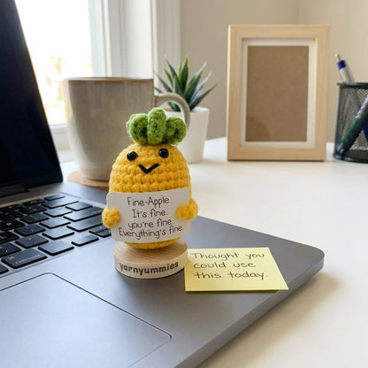 Crocheted pineapple plush decor with a message on a desk next to a laptop