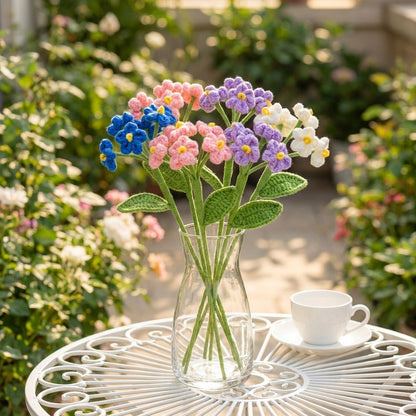 Bouquet of colorful knitted flowers in a clear vase on a garden table with a cup.