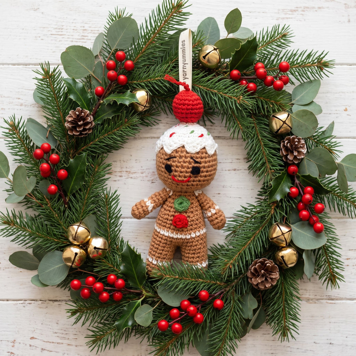 Handmade crochet baby safe gingerbread man ornament by YarnYummies front view showing icing details and embroidered eye and smile hangs from a green pine wreath decorated with red berries, gold bells, pinecones, and eucalyptus leaves on a white wood background.