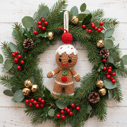 Handmade crochet baby safe gingerbread man ornament by YarnYummies front view showing icing details and embroidered eye and smile hangs from a green pine wreath decorated with red berries, gold bells, pinecones, and eucalyptus leaves on a white wood background.