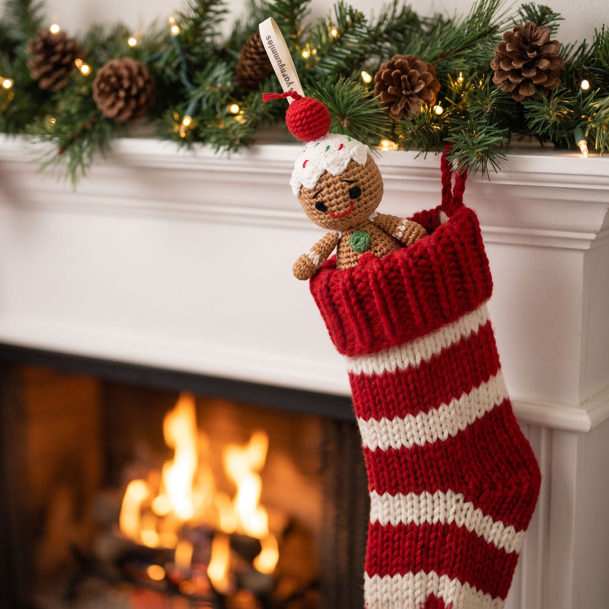 A red and white striped knitted Christmas stocking hangs from a white fireplace mantel decorated with pinecones, evergreen branches, and warm fairy lights. A Handmade crochet baby safe gingerbread man ornament by YarnYummies front view showing icing details and embroidered eye and smile  is tucked inside the stocking, and a warm fire burns in the blurred fireplace behind it.