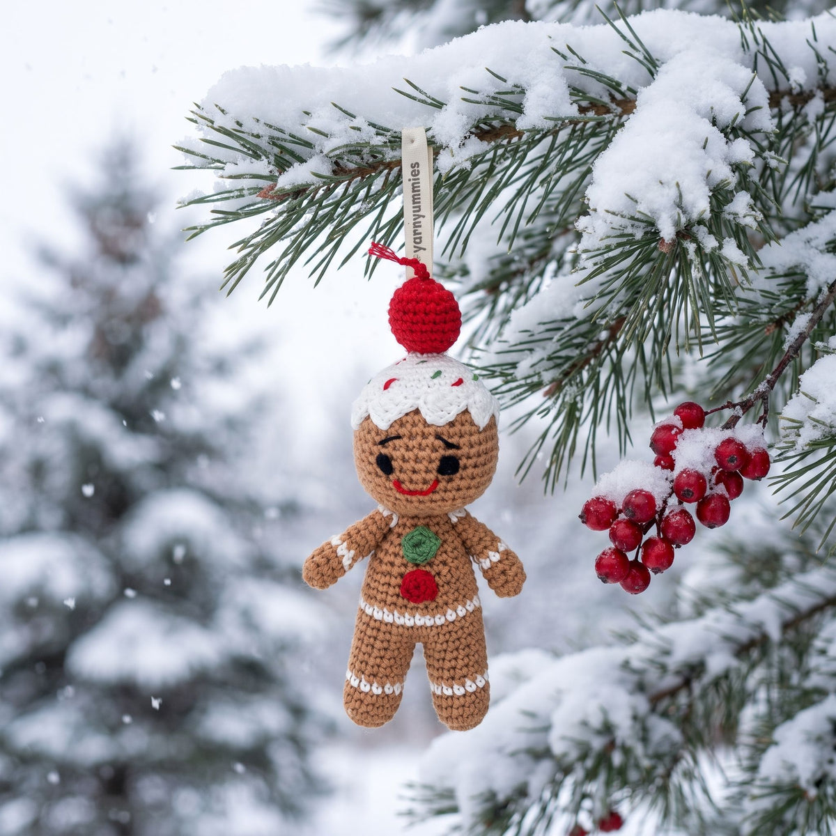 A Handmade crochet baby safe gingerbread man ornament by YarnYummies front view showing icing details and embroidered eye and smile  hangs from a snow-covered evergreen branch. The ornament is tan with white icing details, a red cherry on top, and a green and red chest decoration, set against a snowy outdoor background with blurred pine trees and red berries.