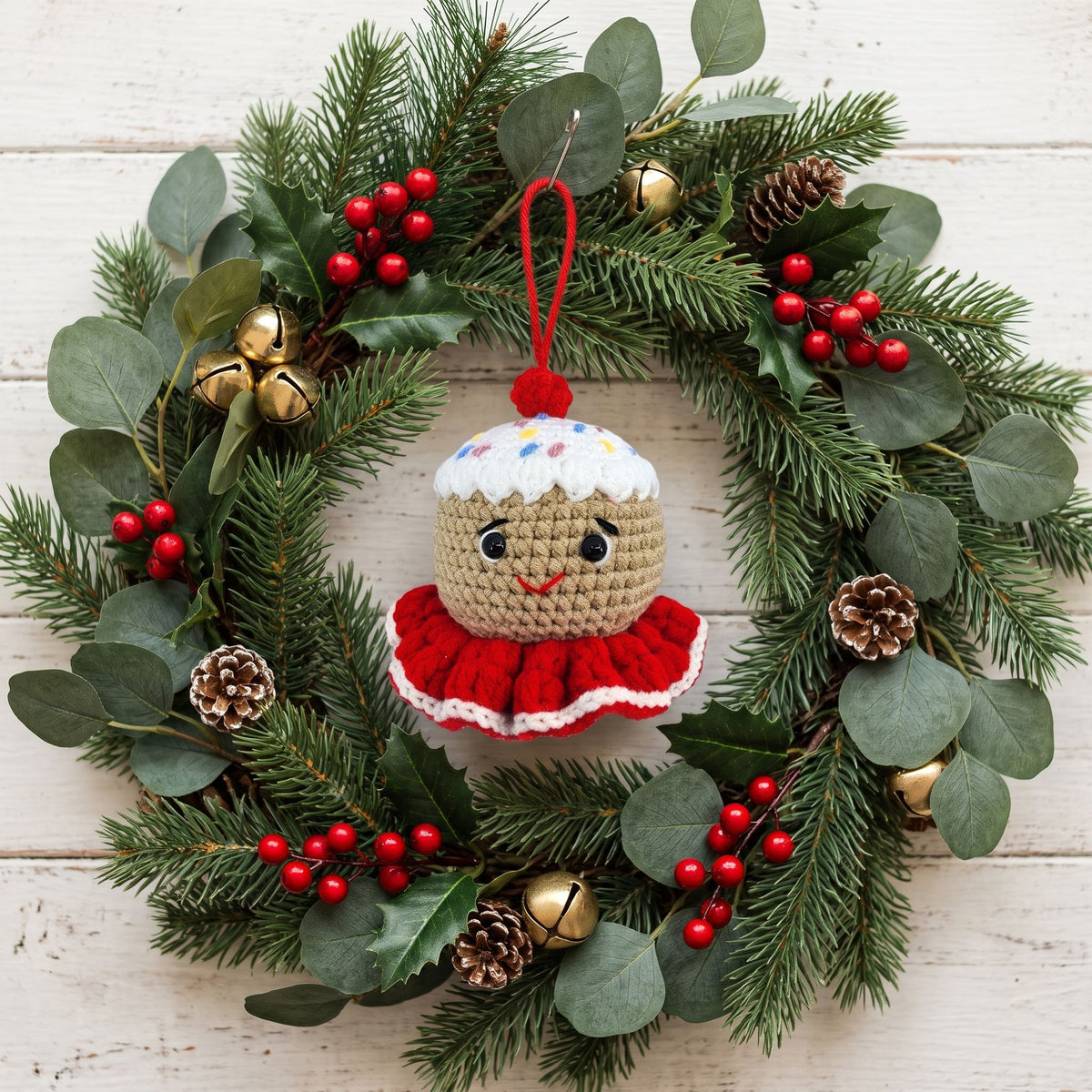 A Handmade crochet gingerbread head ornament by YarnYummies front view showing icing details and sprinkles hanging from a green pine wreath decorated with eucalyptus leaves, red berries, pinecones, and small gold bells on a white wood plank background.
