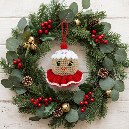 A Handmade crochet gingerbread head ornament by YarnYummies front view showing icing details and sprinkles hanging from a green pine wreath decorated with eucalyptus leaves, red berries, pinecones, and small gold bells on a white wood plank background.