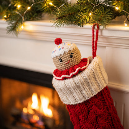A red and cream knitted Christmas stocking with a Handmade crochet gingerbread head ornament by YarnYummies front view showing icing details and sprinkles peeking out and hanging from a fireplace mantel decorated with pine branches and warm string lights.