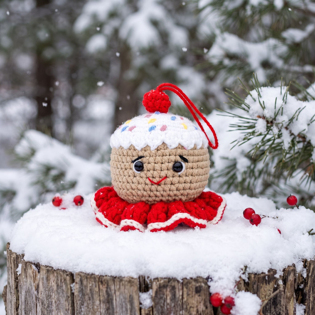 A Handmade crochet gingerbread head ornament by YarnYummies front view showing icing details and sprinkles and a smiling face, placed on a snowy log outdoors with snow-covered pine branches in the background.