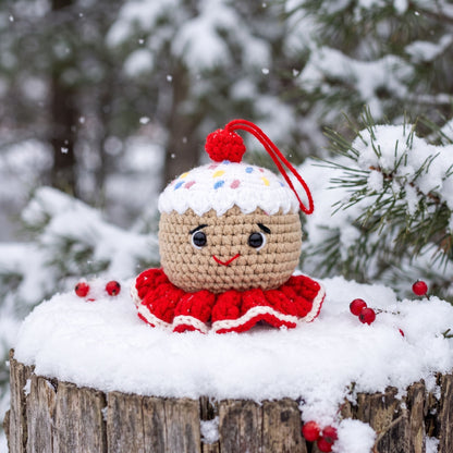 A Handmade crochet gingerbread head ornament by YarnYummies front view showing icing details and sprinkles and a smiling face, placed on a snowy log outdoors with snow-covered pine branches in the background.