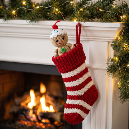A red and white striped knitted Christmas stocking hangs from a white fireplace mantel with a warm fire burning below and holiday greenery with string lights above. Handmade crochet full body gingerbread man ornament by YarnYummies front view showing candy buttons and icing details sits inside the stocking.