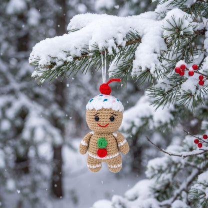 A Handmade crochet full body gingerbread man ornament by YarnYummies front view showing candy buttons and icing details hanging from a snow-covered evergreen branch. The ornament is tan with white icing details, a red pom-pom on top, a green button, and a red button on the front.