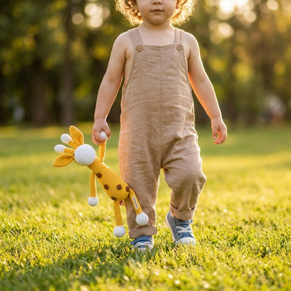 Child in beige overalls holding a giraffe toy in a grassy field