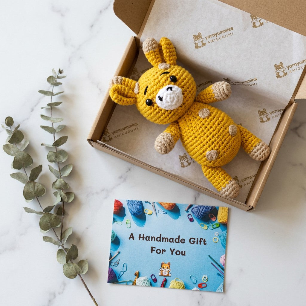 Yellow crochet giraffe in a cardboard box with a 'Handmade Gift' card on a marble surface.