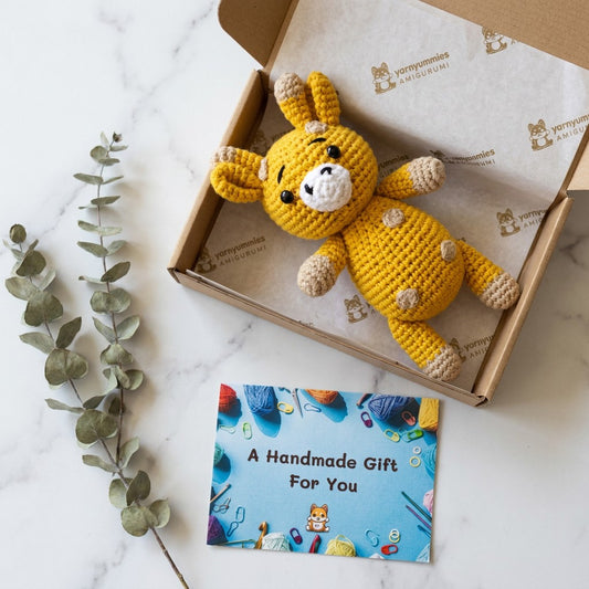 Yellow crochet giraffe in a cardboard box with a 'Handmade Gift' card on a marble surface.