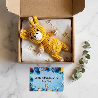 Yellow crocheted giraffe toy in a box with a card on a marble surface