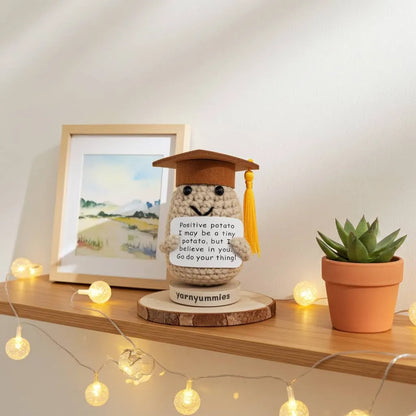 Crocheted positive potato character plush with a graduation cap on a shelf with decorative lights and a plant.