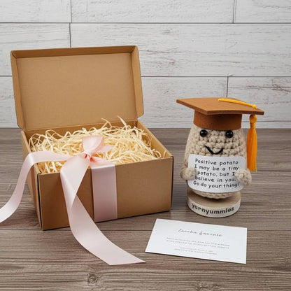 Graduation-themed crocheted positive potato character plush toy with a message card next to an open gift box on a wooden surface.