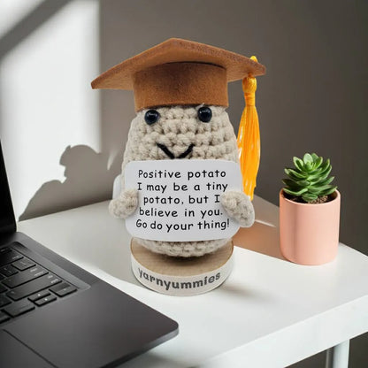 Graduation-themed crocheted positive potato character plush with a motivational quote on a desk next to a laptop and plant.