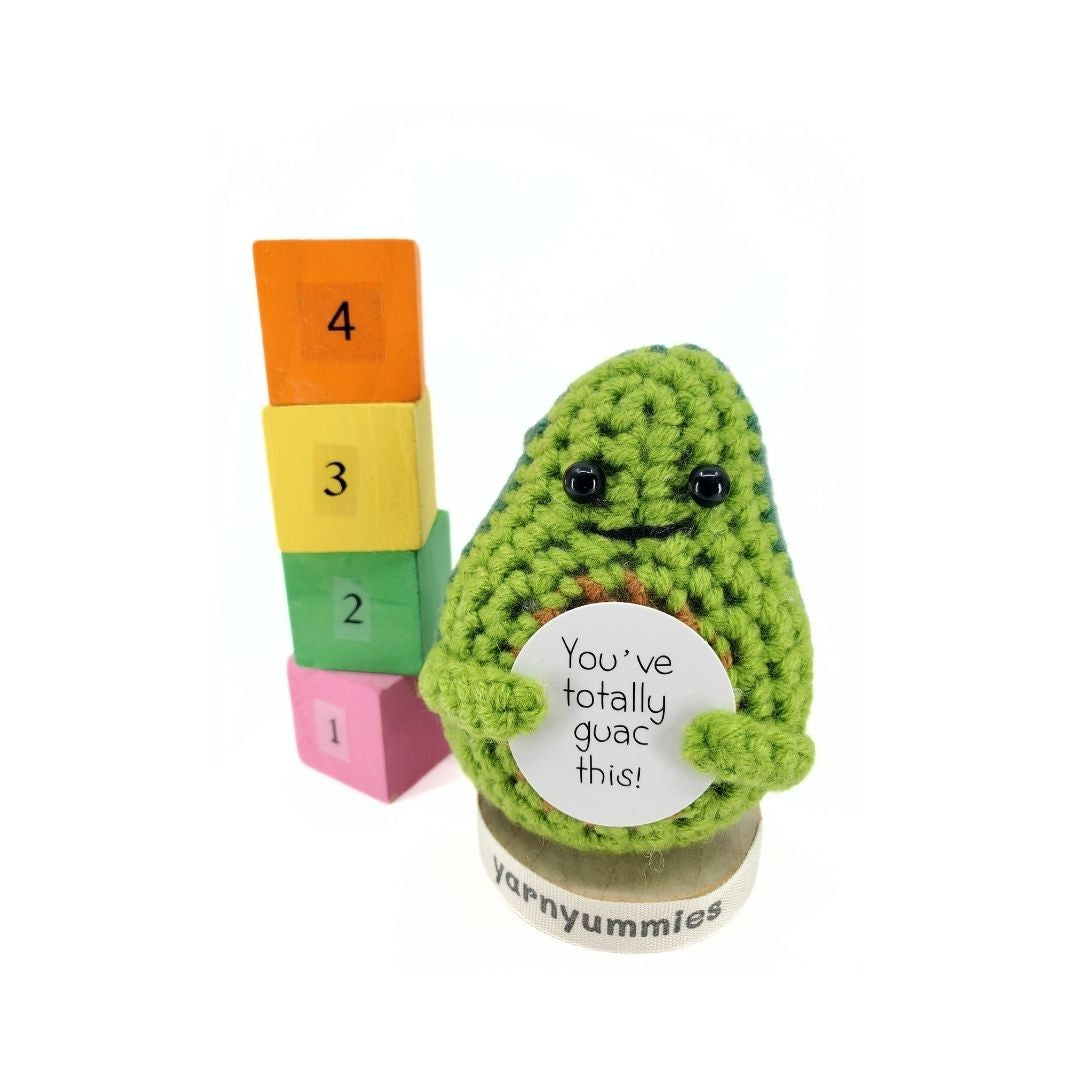 Crocheted avocado plush decor with a message tag next to colorful building blocks on a white background