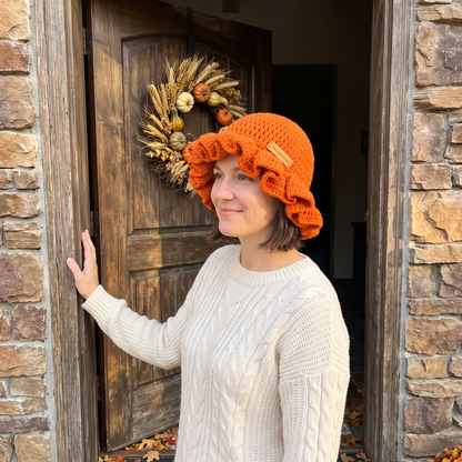 Woman wearing an orange handmade crochet ruffled brim bucket hat by YarnYummies standing at a rustic front door with a fall harvest wreath