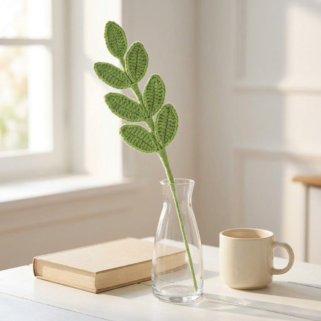 Crocheted leafy branch in a clear vase on a table with a book and mug.