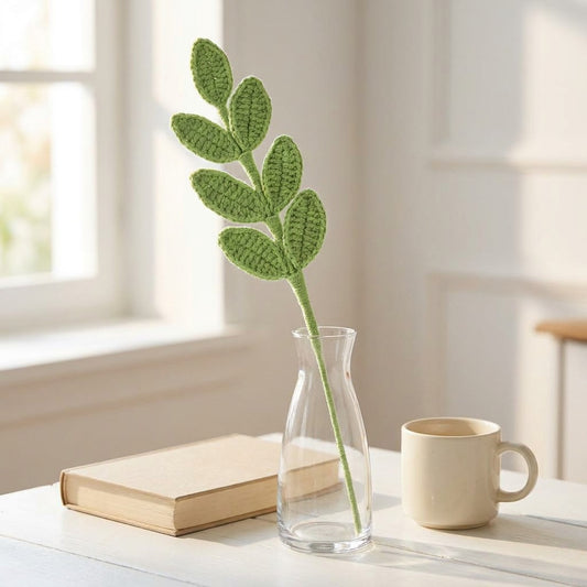 Crocheted leafy branch in a clear vase on a table with a book and mug.