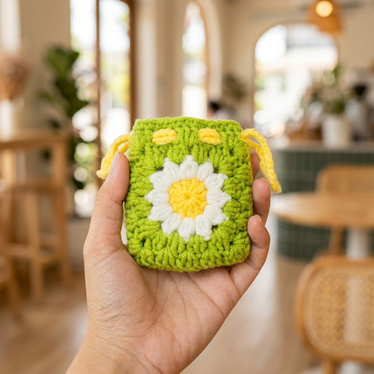 Hand holding a green crocheted pouch with a white flower design in a blurred indoor setting