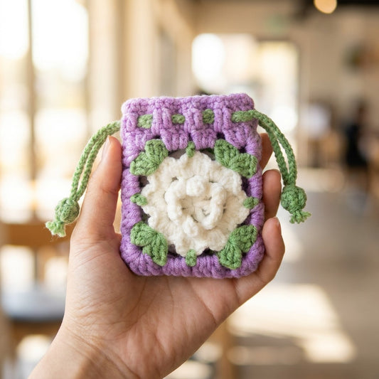 Hand holding a purple and green crocheted pouch with a white floral design.