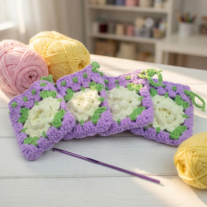 Crocheted blanket with purple and white flowers, surrounded by yarn and a crochet hook on a wooden surface.