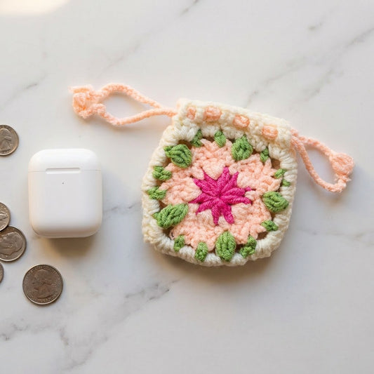 Crocheted pouch with pink center and green leaves on a marble surface with coins and an airpods case.