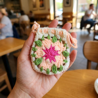 Hand holding a crocheted square with pink center and green leaves in a casual setting.