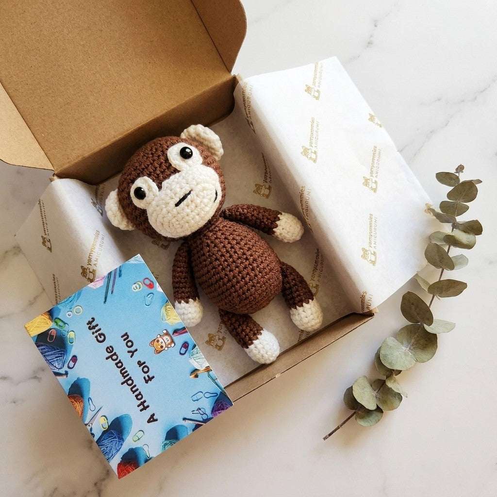 Crocheted monkey toy in an open box with a handkerchief and eucalyptus leaves on a marble surface.