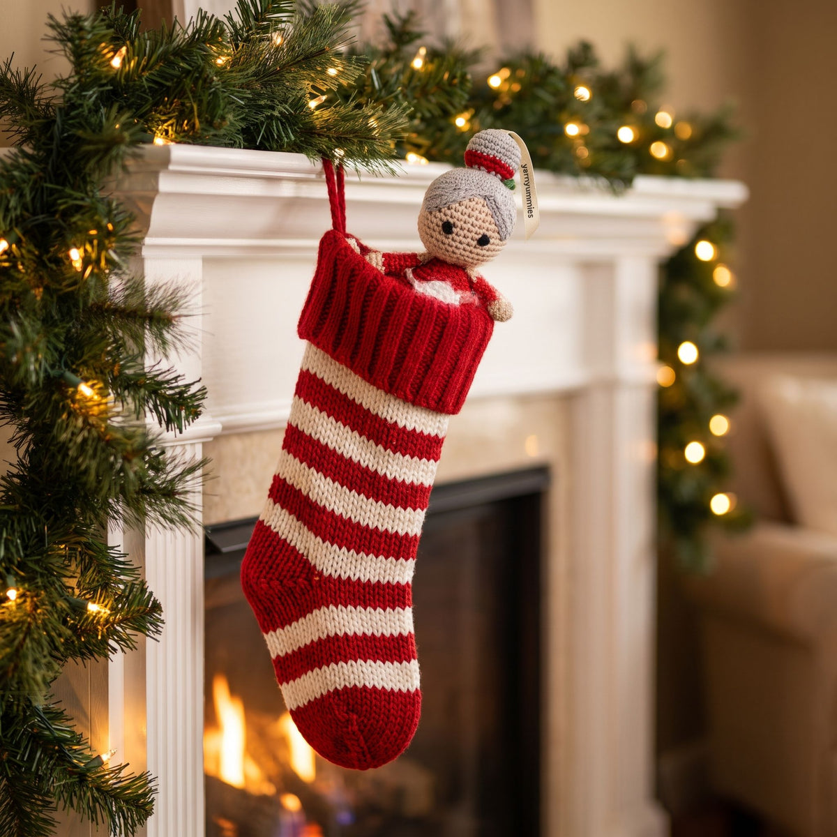 A red and cream striped knit Christmas stocking with a handmade crochet baby safe Mrs. Claus ornament by YarnYummies hanging from a white fireplace mantel decorated with green garland and warm string lights.